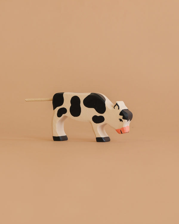 The Holztiger Calf, Grazing is a small handcrafted wooden toy with black and white spots, standing on a light brown surface against a beige background—showcasing the European craftsmanship of HOLZTIGER figures.