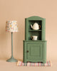 A dark green Maileg Large Corner Cabinet with a vintage finish displays a white teapot and teacup on its shelves, standing on a striped rug. Next to it is a light blue floor lamp with a floral patterned lampshade. The backdrop is a beige wall, creating stylish home decoration reminiscent of miniature house furniture.