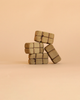 Four small Bumbu Handmade Wooden Haystacks Set packages, wrapped in brown paper and tied with string, are stacked on a beige background with one leaning to the side.