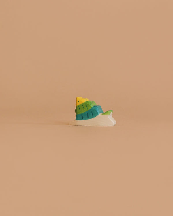 A small, colorful snail with layers of yellow, green, blue, and white—handcrafted in Europe and inspired by HOLZTIGER figures—sits on a plain beige background. This is the Holztiger Snail wooden toy.