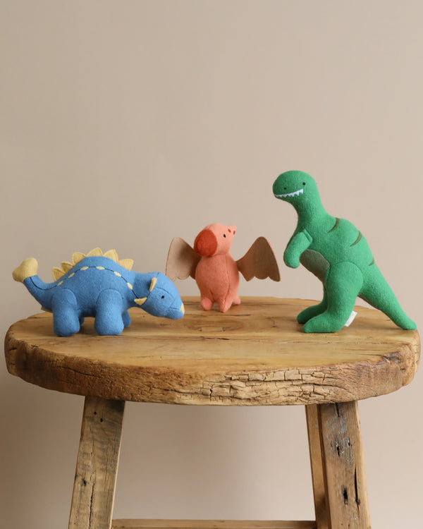 Three Olli Ella Holdie Pre-Historic Animals—a blue stegosaurus, a pink triceratops, and a green T-Rex—standing on a rustic wooden table.