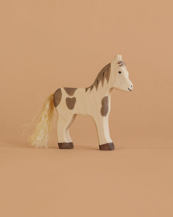 The Holztiger Foal, Standing is a handcrafted wooden toy horse with painted brown patches, dark brown hooves, and a light-colored string mane and tail, displayed on a plain beige background.