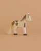 The Holztiger Foal, Standing is a handcrafted wooden toy horse with painted brown patches, dark brown hooves, and a light-colored string mane and tail, displayed on a plain beige background.