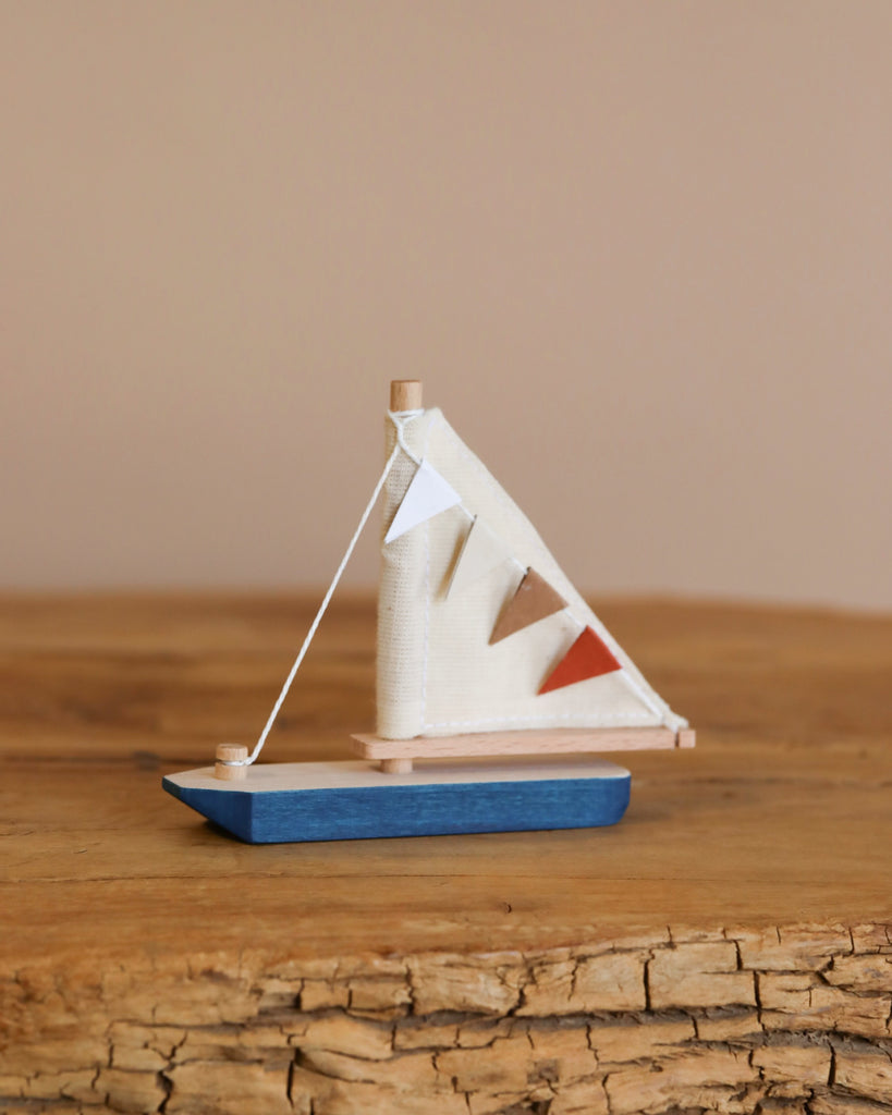 The Handmade Small Wooden Boat from Ukraine features a blue hull on wood, with a white sail decorated with three triangular flags in white, blue, and orange. It’s painted with non-toxic paint and set against a plain beige background.