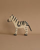 A wooden zebra figure stands against a beige background. The Holztiger Zebra has black stripes and a simple, minimalist design, with a flat body and a tail. This high-quality, made-in-Europe piece showcases the artistry found in handcrafted wood toys.