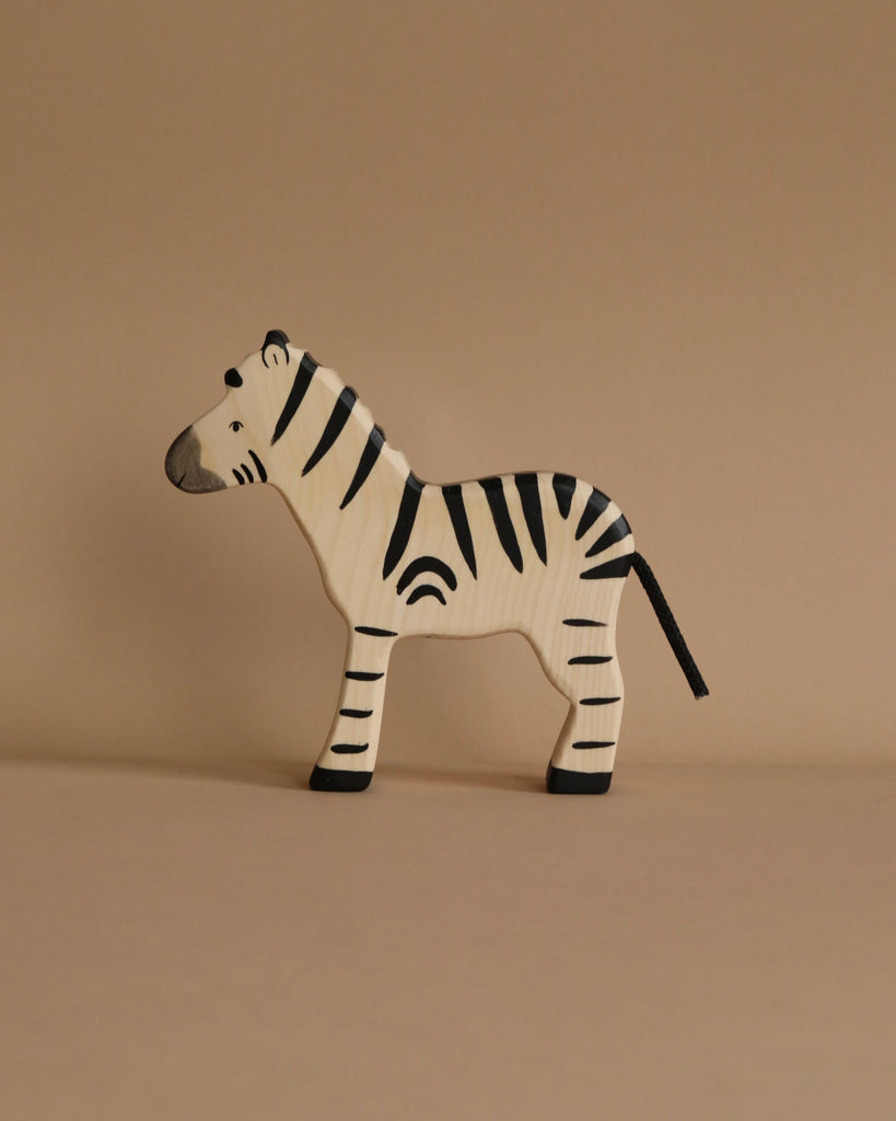 A handcrafted zebra from the Holztiger Handmade Wooden Safari Animals Set, featuring black stripes, stands on a beige surface—ideal for imaginative play and a charming addition to any Holztiger collection.