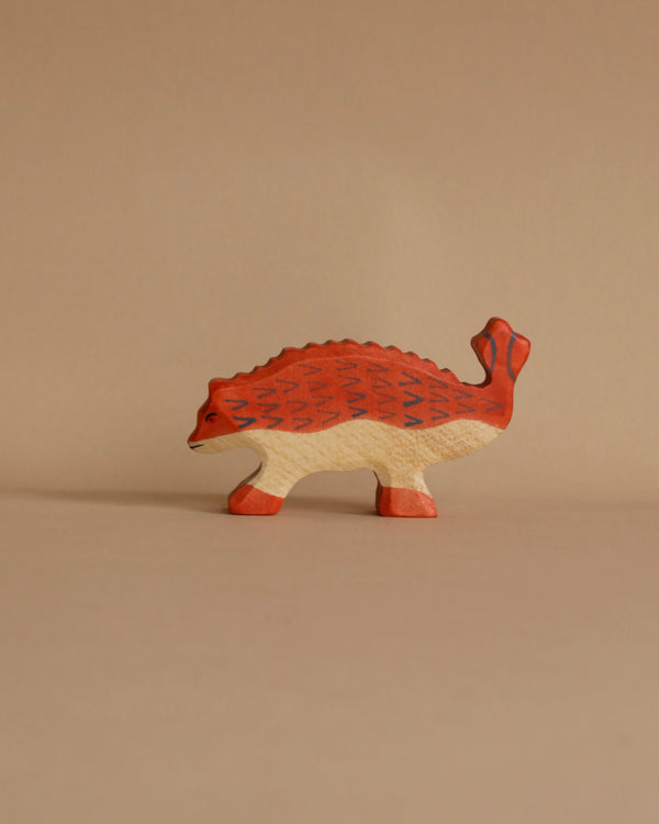 A small, hand-carved wooden toy in the shape of an animal, painted in shades of orange and brown with patterned markings along its body. This Holztiger Ankylosaurus Dinosaur has four legs, a tail lifted upwards, and a pointed snout, reminiscent of HOLZTIGER figures, set against a plain beige background.