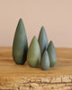 The Handmade Wooden Bush Set features five cone-shaped wooden figures in different sizes and green shades, crafted with non-toxic paint. Arranged on wood against a neutral backdrop, these charming tree-like pieces are proudly made in Ukraine.