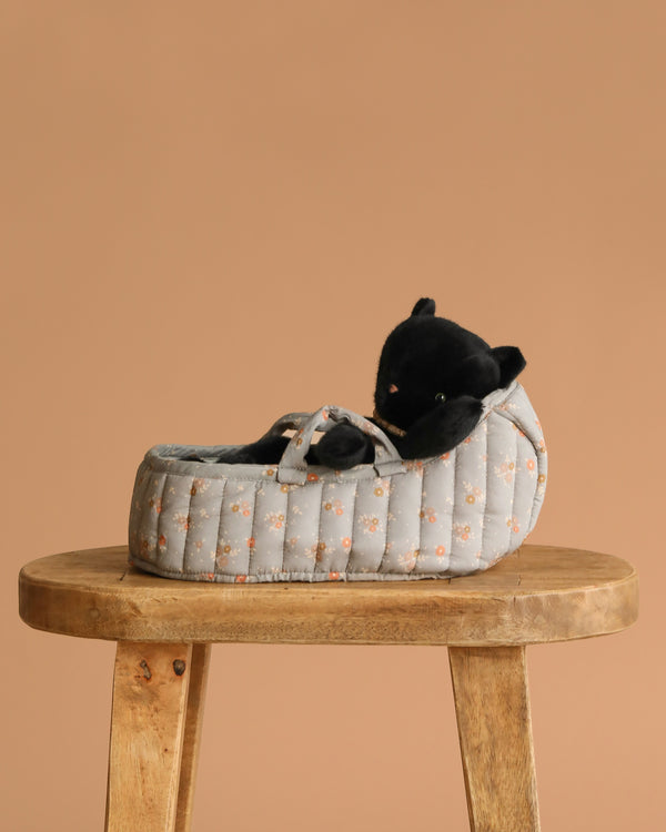 The Maileg Sleeping Kitten in Carry Cot features a plush kitten nestled in a quilted gray and orange floral-patterned carry cot, displayed on a round wooden stool against a plain beige background.