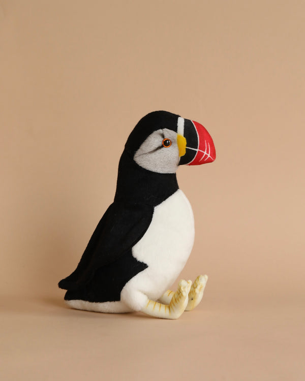 A Puffin Bird Stuffed Animal with a black and white body and colorful beak, sitting against a plain, light brown background. This realistic plush animal captures the charming essence of its wild counterpart.