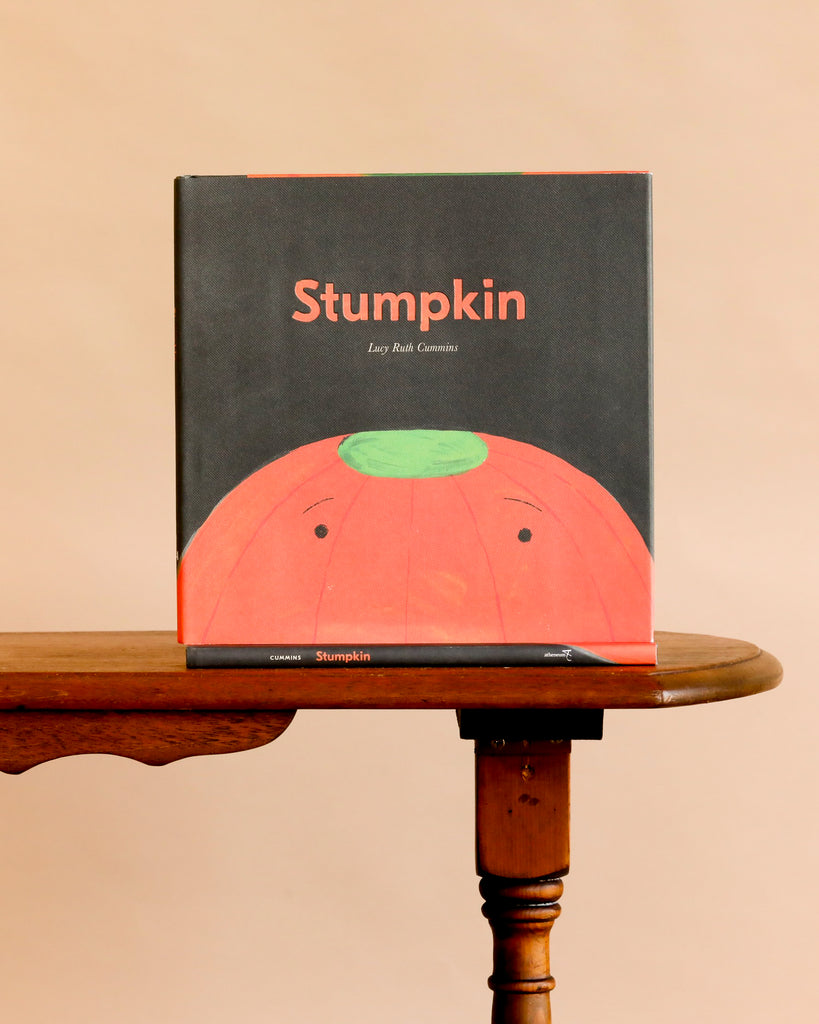 Stumpkin by Lucy Ruth Cummins, a children’s Halloween pumpkin book, stands upright on a wooden table; its cover features Stumpkin—a stemless jack-o-lantern with a green top on a beige background.