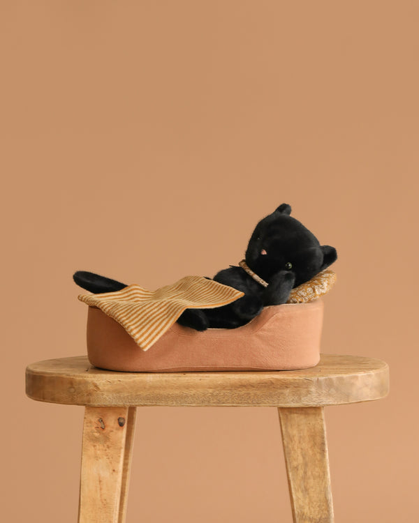 A Maileg Sleeping Kitten in a cozy bed with a yellow striped pillow and blanket rests on a wooden stool against a beige background.