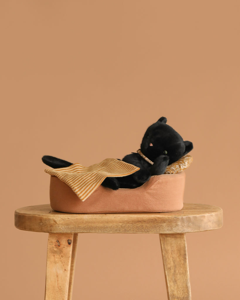 A Maileg Sleeping Kitten in a cozy bed with a yellow striped pillow and blanket rests on a wooden stool against a beige background.