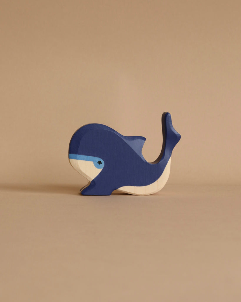 A small, blue wooden whale from the Holztiger Handmade Wooden Ocean Animals Set with painted details rests on a light brown surface against a matching background, evoking classic handcrafted toy charm.