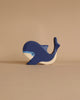 A small wooden toy whale painted in shades of blue and white is placed against a plain beige background. This Holztiger Blue Whale figure has a simple, stylized design with a friendly appearance. Its painted eye and curved tail add to its charm, showcasing the artistry of handcrafted wood toys made in Europe.