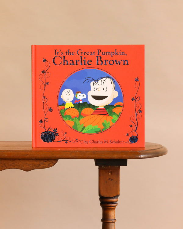 A copy of It’s the Great Pumpkin, Charlie Brown by Charles M. Schulz stands on a wooden table, featuring Peanuts characters and pumpkins on its red cover.