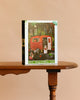 The Dogs of Halloween - 500 Piece Jigsaw Puzzle sits on a wooden table, featuring an illustration of costumed dogs near a red trailer amid Halloween decorations.