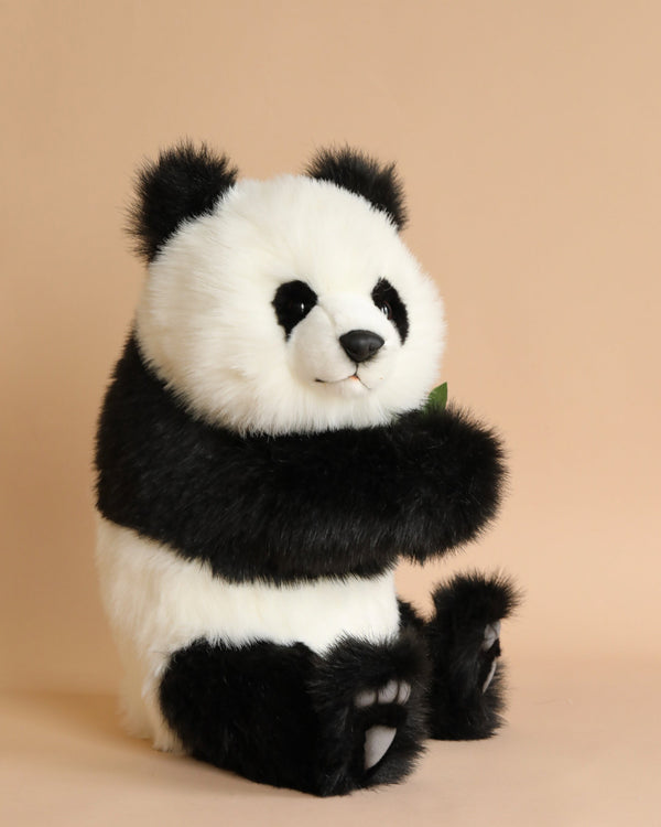 A Panda Cub Stuffed Animal, hand sewn with black and white fur, sitting upright against a plain beige background, holding a small green leaf. Its eyes are round and shiny.