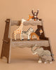 A wooden step stool displays the Holztiger Handmade Wooden Safari Animals Set—giraffe, zebra, lion, elephant with calf, tiger, and chimpanzee—against a light brown background.