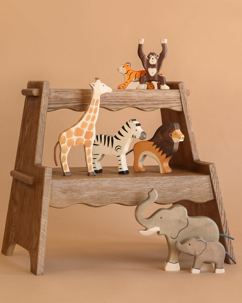 A wooden step stool displays the Holztiger Handmade Wooden Safari Animals Set—giraffe, zebra, lion, elephant with calf, tiger, and chimpanzee—against a light brown background.