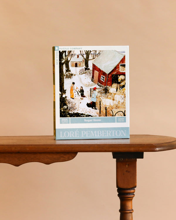 A cozy farm puzzle with snowy village art stands on a wooden table against a beige background. The box reads “Sugar Snow - 500 Piece Jigsaw Puzzle” by Loré Pemberton.