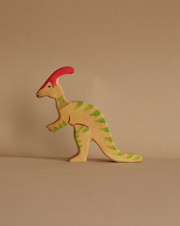 A handcrafted wooden toy dinosaur figurine stands on a plain surface. The Holztiger Parasaurolophus Dinosaur, made in Europe, is painted light green with pink ridges along its back and a red crest on its head, set against a neutral beige background.