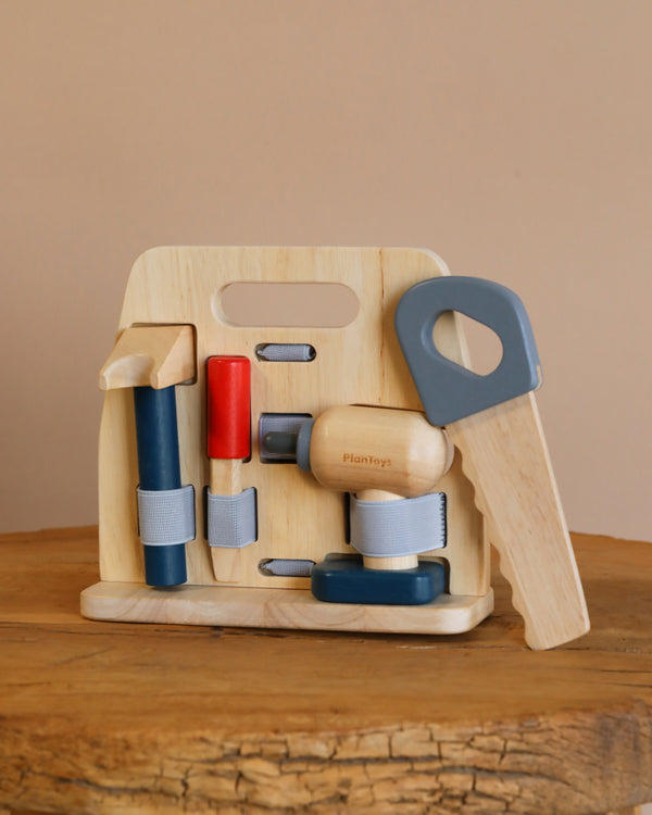 The Handy Carpenter Tool Set rests on a wooden surface, showcasing a claw hammer, saw, screwdriver, wrench, and a red-tipped tool. All tools are neatly secured with gray elastic straps on the wooden stand.