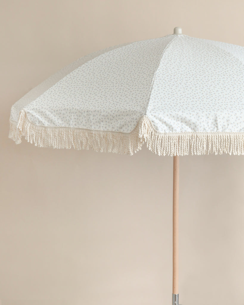 A beige Beach Umbrella with a white paisley pattern and fringe details, positioned upright against a neutral-toned background.
