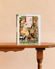 Quill and Parchment - 1000 Piece Jigsaw Puzzle rests on a wooden table, showing an illustrated scene of two people in a cozy bookstore filled with bookshelves and a large window.