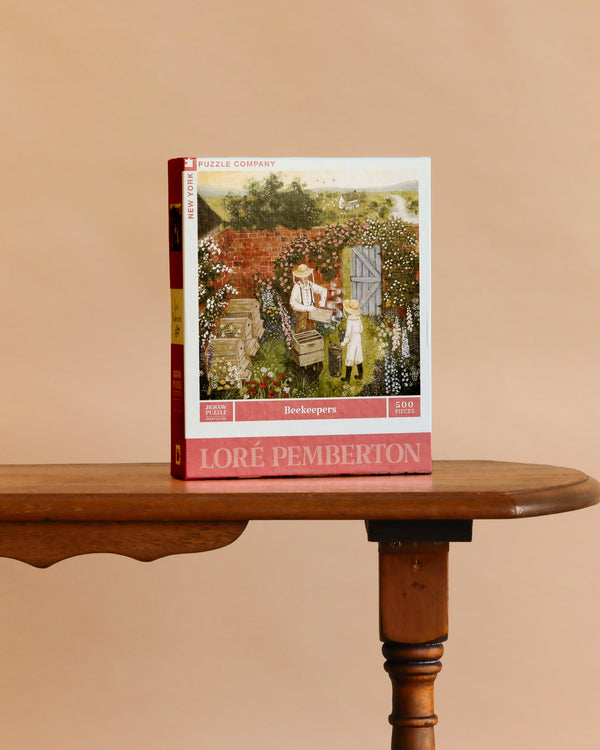A box of the Beekeepers - 500 Piece Jigsaw Puzzle stands upright on a wooden table against a plain beige background, showcasing an illustration of beekeepers at work.