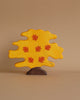 A handcrafted wood tree figure with a bright yellow canopy and orange leaves. The dark brown base complements its whimsical, playful appearance, typical of Holztiger Maple Tree figures. Set against a plain beige background, this charming piece adds delightful play value to any setting.