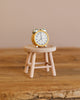 A small yellow alarm clock sits on a light wooden stool atop a rustic wooden surface with a neutral beige background, creating the perfect setting for the Maileg Cozy Christmas Corner.