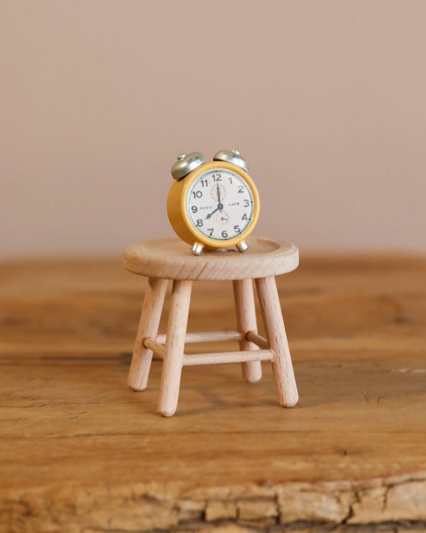 A Maileg Alarm Clock For Dollhouse in yellow sits on a tiny wooden stool against a blurred, warm-toned wall, creating a cozy atmosphere. The clock's hands display 7:10, enhancing its whimsical charm.