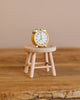 A Maileg Alarm Clock For Dollhouse in yellow sits on a tiny wooden stool against a blurred, warm-toned wall, creating a cozy atmosphere. The clock's hands display 7:10, enhancing its whimsical charm.