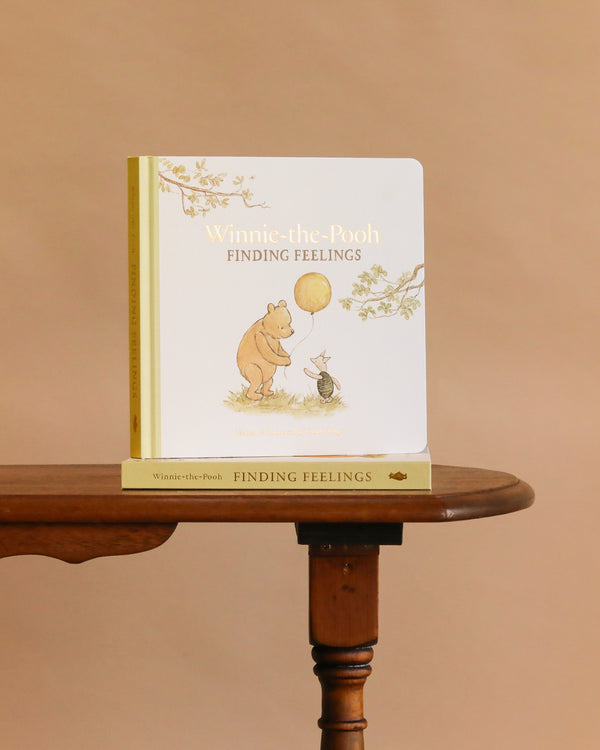 A copy of “Winnie-the-Pooh: Finding Feelings” stands upright on a small wooden table with another copy below, set against a plain beige background.