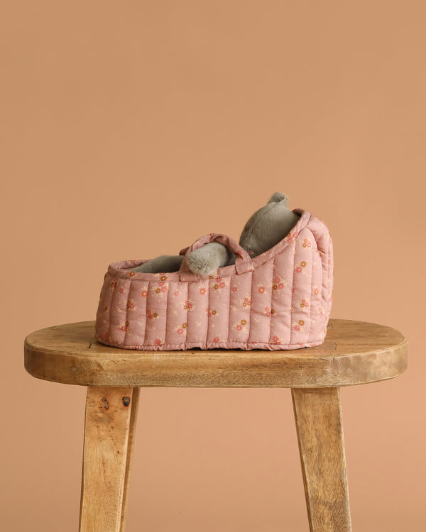 The Maileg Sleeping Kitten in Carry Cot rests beside a small pink floral baby shoe with gray lining on a wooden stool, set against a plain beige background.