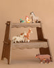 The Holztiger Handmade Wooden Farm Animals Set, crafted from maple and beech wood, features a duck, chicken, donkey, goat, horse, dog, pig, and cow on a wooden display shelf against a plain beige background.