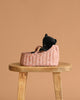 A Maileg Sleeping Kitten in a pink, patterned carry cot rests on a wooden stool against a plain beige background.
