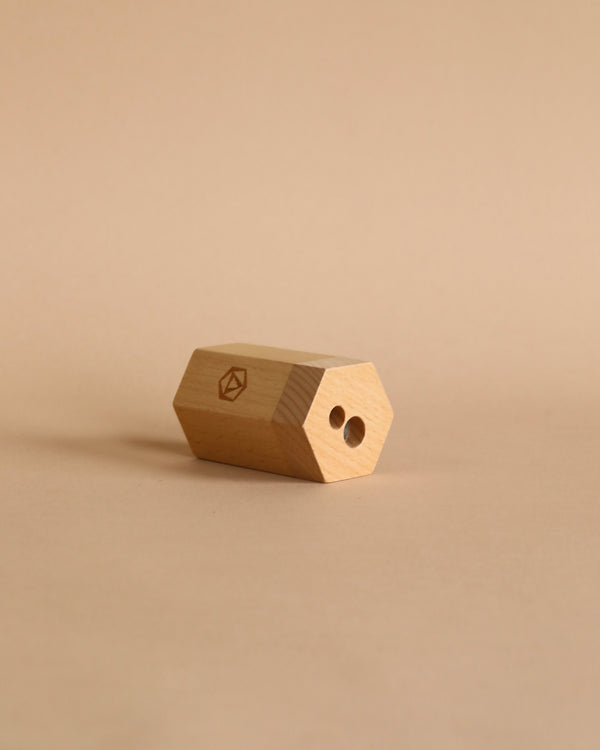 A Stockmar Dual Pencil Sharpener with a hexagonal shape and two holes, made from FSC certified lime wood, placed on a plain beige background. The sharpener has a small engraved symbol on one side.