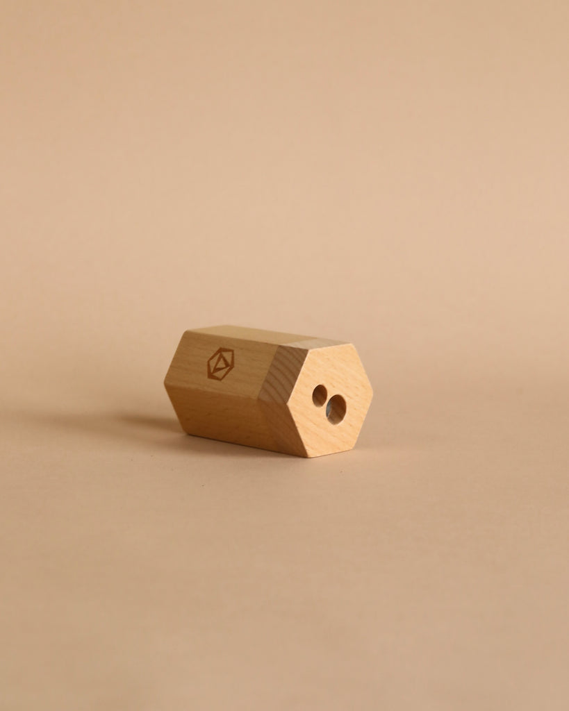 A hexagonal wooden dual pencil sharpener with two holes sits on a beige surface, sporting a small geometric logo—ideal for use with the Moulin Roty Botanist Coloring Book with Stockmar Pencil Set.