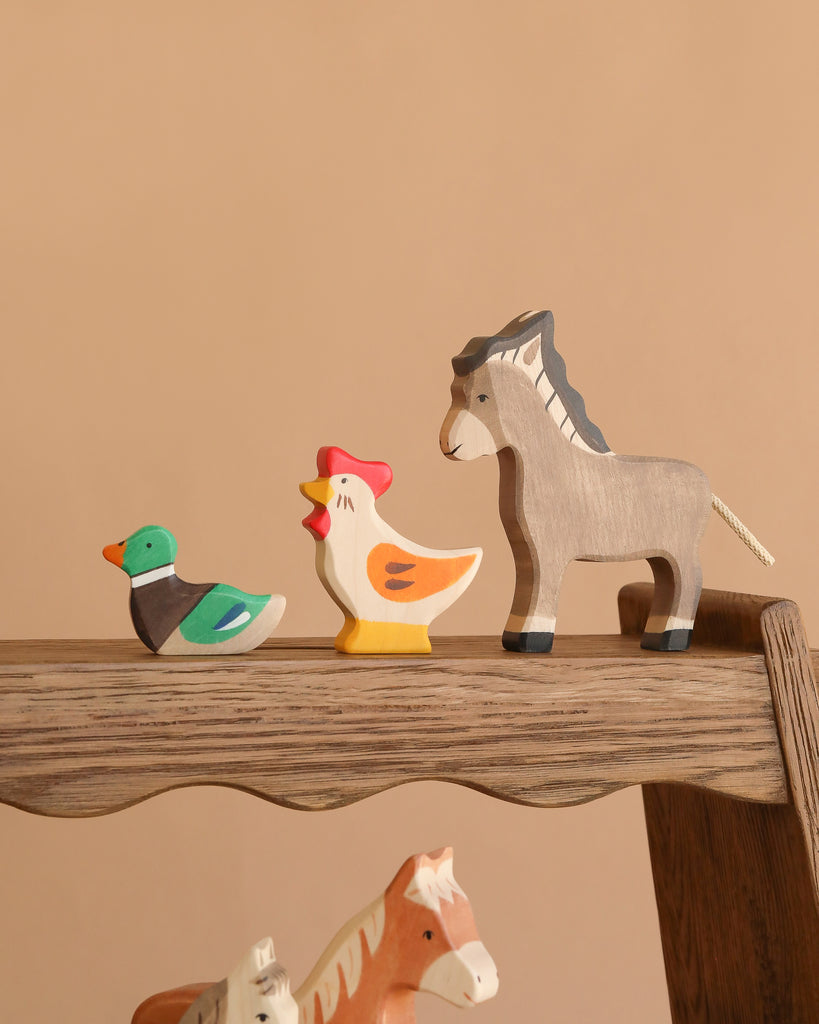 The Holztiger Handmade Wooden Farm Animals Set, featuring a green and brown duck, a white and yellow chicken, and a gray donkey crafted from maple and beech wood, is displayed in a row on a wooden shelf against a beige background.