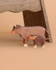 Two figures from the Holztiger Handmade Wooden Farm Animals Set—a cow and a calf crafted from maple and beech wood—stand side by side on a tan surface, with a wooden chair leg visible in the background.