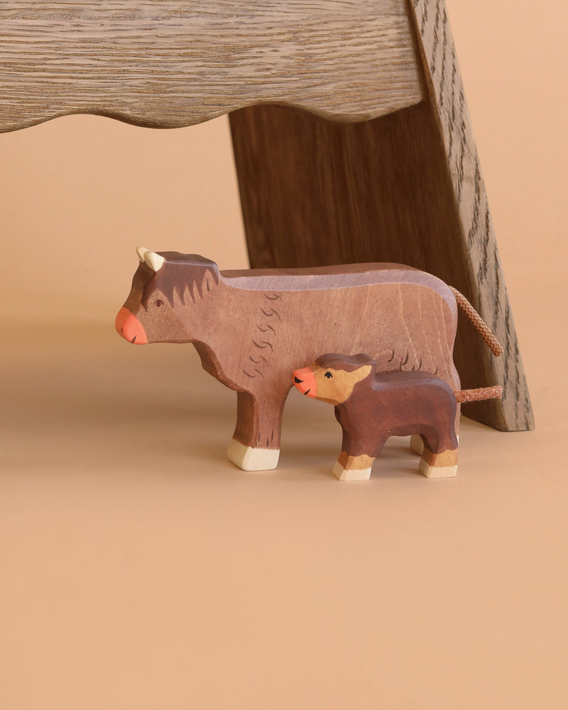 Two figures from the Holztiger Handmade Wooden Farm Animals Set—a cow and a calf crafted from maple and beech wood—stand side by side on a tan surface, with a wooden chair leg visible in the background.