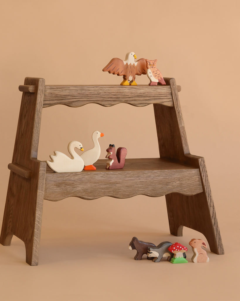 A small wooden display stand with two shelves holds the Holztiger Handmade Wooden Woodland Animals Set, featuring figures like an eagle, owl, swans, and squirrel. More handmade wooden toys and mushroom figures are arranged on the floor below.