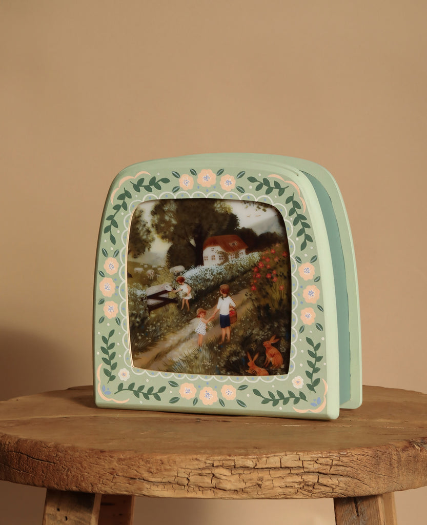 The vintage-style photo frame captures children in a garden with a quaint cottage, while the Hand-Painted Toverlux Magic Lamp - Green Meadow casts a soft glow on the rustic wooden surface against a beige wall, creating cozy silhouettes.