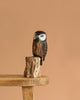 The Hand Carved Ornament - Saw Whet Owl - 10"H features a painted wooden owl with lifelike eyes, perched on a small tree stump atop a wooden bench, set against a plain light brown background.