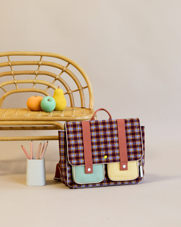A stylish Sticky Lemon School Bag from the Adventure Collection in the Tartan Stormy Purple color sits in front of a bamboo bench. On the bench are colorful fruit-shaped toys and a container holding art supplies. The bag is made from waterproof materials.