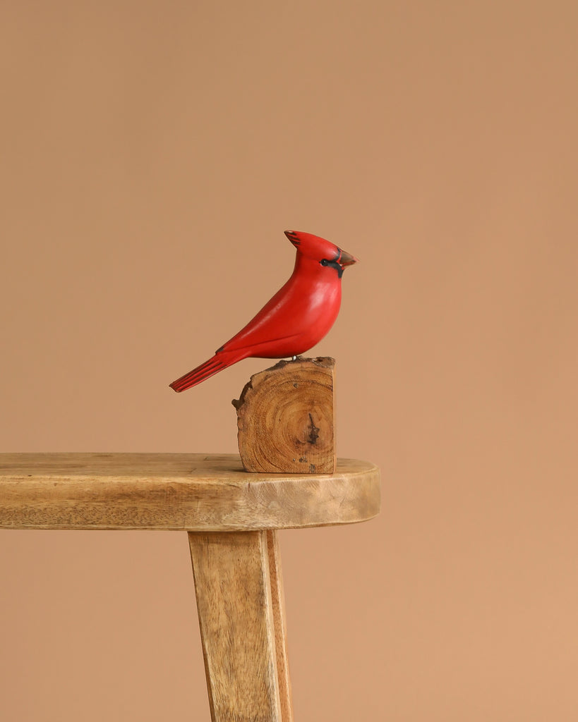 The Hand Carved Ornament - Cardinal Bird - Male - 7"H features a handmade wooden red cardinal figurine perched on wood atop a bench, creating a charming decorative accent against a beige background.