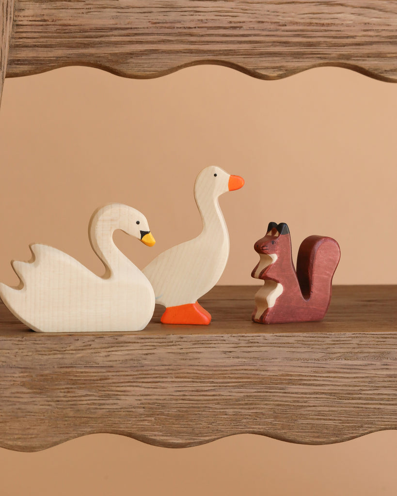 Part of the Holztiger Handmade Wooden Woodland Animals Set, a white swan, a white duck with orange beak and feet, and a brown squirrel made from maple and beech wood stand on a wooden shelf against a beige background.