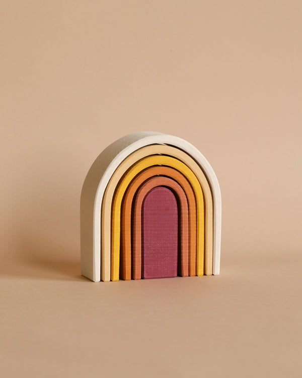 A colorful Raduga Grez Handmade Wooden Oval Rainbow Stacker with seven arches in shades of cream, yellow, orange, and pink, stacked neatly together against a pale beige background.